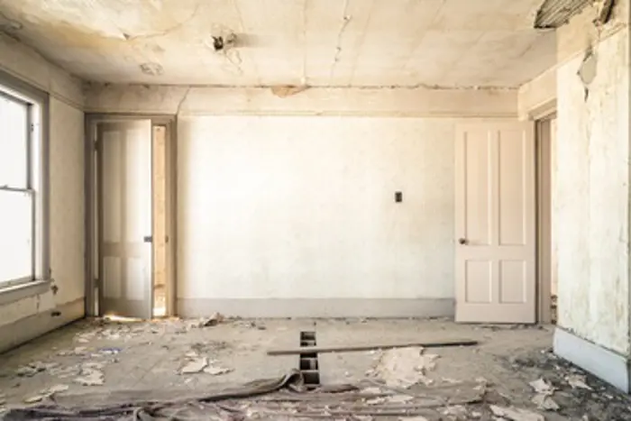 An empty, rundown room with peeling paint on the walls and ceiling, exposed floorboards, and debris scattered on the floor suggests an investigation may reveal underlying issues. Two doors stand open on either side, leading to other rooms. Natural light enters through a single window on the left.