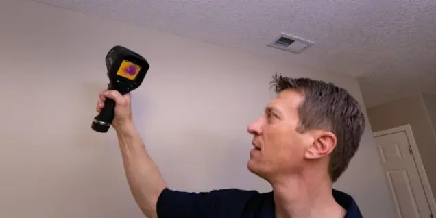 A person holding a thermal imaging camera up to a ceiling vent, conducting an inspection for temperature differences or leaks that could indicate mold or require further sampling.