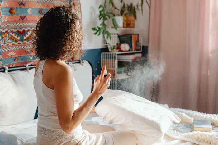 A person with curly hair sits on a bed, carefully inspecting the mist they spray onto a pillow. The room, filled with colorful textiles, plants, and bookshelves in the background, conveys a sense of relaxation and cleanliness.