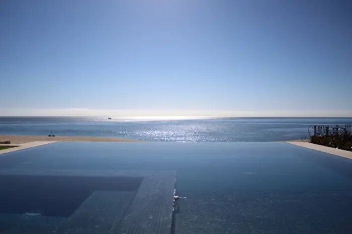 Infinity pool overlooking a calm, sunlit ocean with a clear blue sky. The horizon is visible with the shimmering water reflecting the sunlight, creating a serene and peaceful atmosphere, inviting inspection of nature's tranquility.