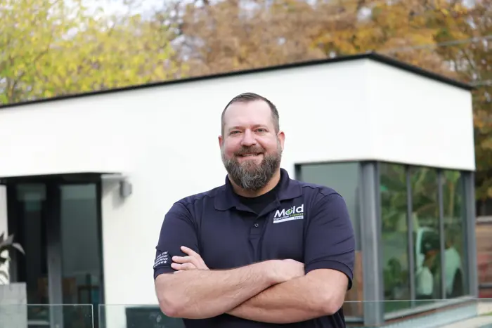 A man with a beard stands confidently with arms crossed, wearing a navy blue shirt that displays the Mold logo. In the background, a modern-style house with large windows and autumn foliage awaits an expert mold assessment to ensure its pristine condition.