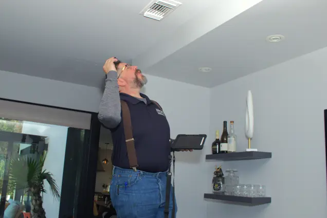 A bearded man stands indoors, conducting a mold inspection with a flashlight. He holds a tablet and wears a dark shirt with suspenders and glasses. On the white wall behind him, shelves brim with bottles and glassware.