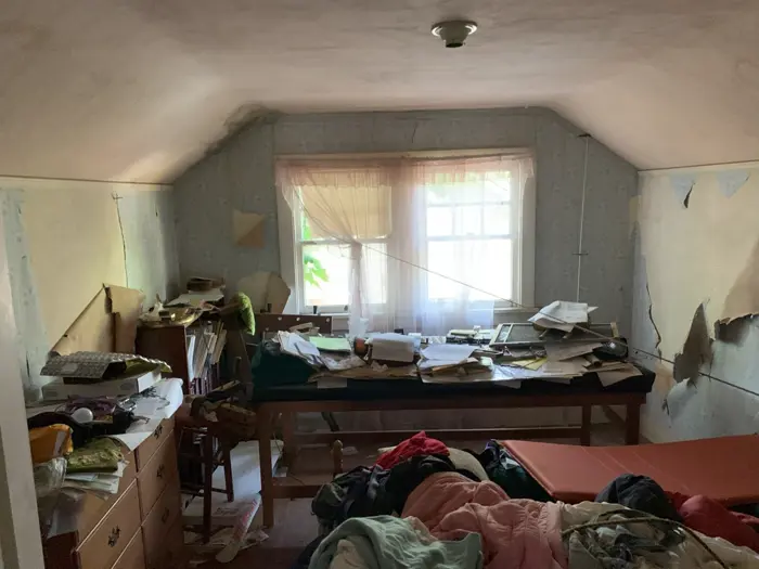 The room, subject to an ongoing investigation, is cluttered with peeling wallpaper and a small window. Papers and various objects pile on the desk. Clothes and a mattress scatter the floor, with drawers and an old chair nearby. The dim lighting adds to the atmosphere of disarray.