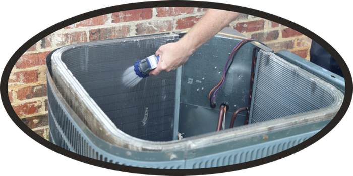 A person uses a hose nozzle to clean the interior coils of an outdoor air conditioning unit near a brick wall, following an inspection to check for mold buildup.