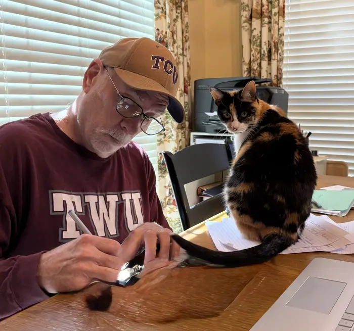 An older man in a TWU shirt and TCU cap conducts a careful inspection of a small object at his desk, while a calico cat sits on papers beside him, looking at the camera as sunlight filters through nearby blinds.