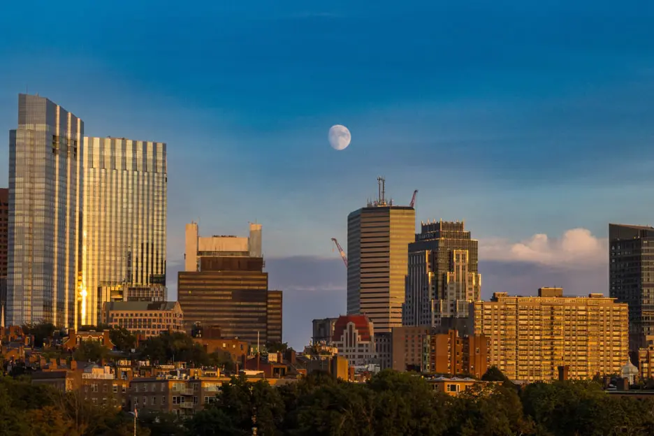 City skyline at sunset with a clear blue sky and a visible moon. High-rise buildings are illuminated by the setting sun, casting a warm glow on the urban landscape, while lush green trees appear in the foreground, creating an impeccable scene that demands further inspection.