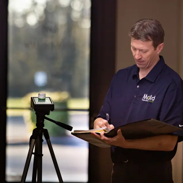 A person wearing a navy Mold shirt is conducting an assessment, writing in a notebook next to a tripod-mounted device. They're standing indoors near a window with blurred greenery outside, focused on their investigation.