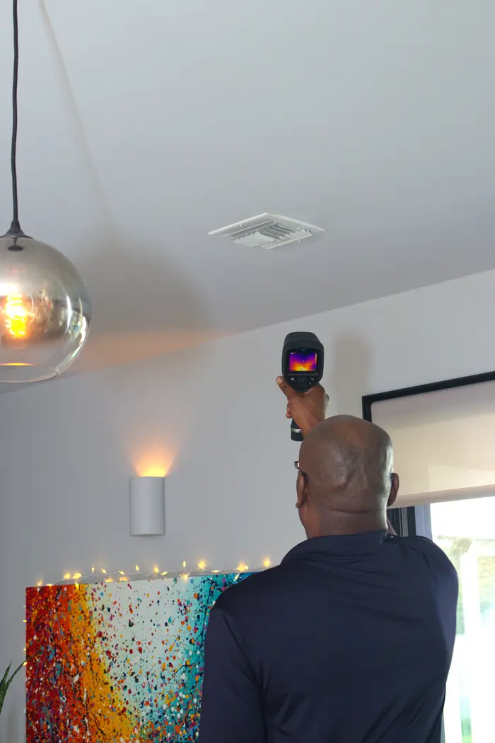 A person conducts an inspection with a thermal imaging camera to assess the ceiling vent. The room is illuminated by a hanging light, with colorful artwork adorning the walls.