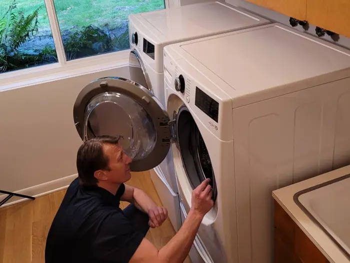 A person kneels in front of a front-loading washing machine with the door open, conducting an inspection of its interior. The room features a window revealing greenery outside and a countertop adjacent to the washer and dryer.