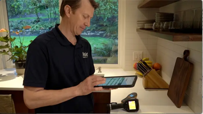 A man in a kitchen uses a tablet to conduct an assessment while standing by the counter. Surrounded by kitchen utensils, a cutting board, and a fruit bowl, he glances at the greenery outside through the window. His dark polo shirt with a logo adds to his focused demeanor.
