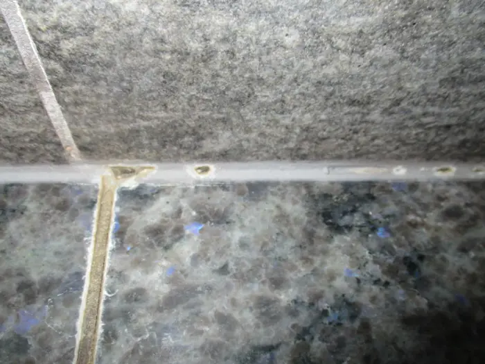 A close-up of a tiled wall and countertop corner reveals gray stone tiles. Upon inspection, there are small holes along the grout line where the wall and countertop meet, with slightly discolored grout.