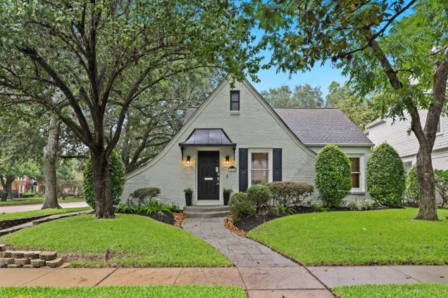 Charming cottage-style house with a steeply pitched roof and light-colored brick exterior. Surrounded by lush green grass, trimmed shrubs, and large trees. A curved walkway leads to a black front door with a small overhang, perfect for investigation of its cozy moldings and unique architectural details.