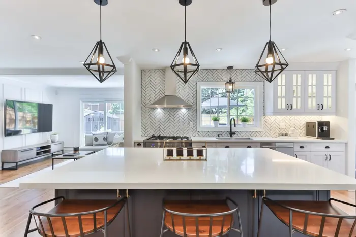 Modern kitchen with a large white island, wooden barstools, and pendant lights complementing the herringbone tile backsplash. A window above the sink offers natural light, perfect for an easy morning assessment of your day. The room extends into a bright living area with a TV and couch.