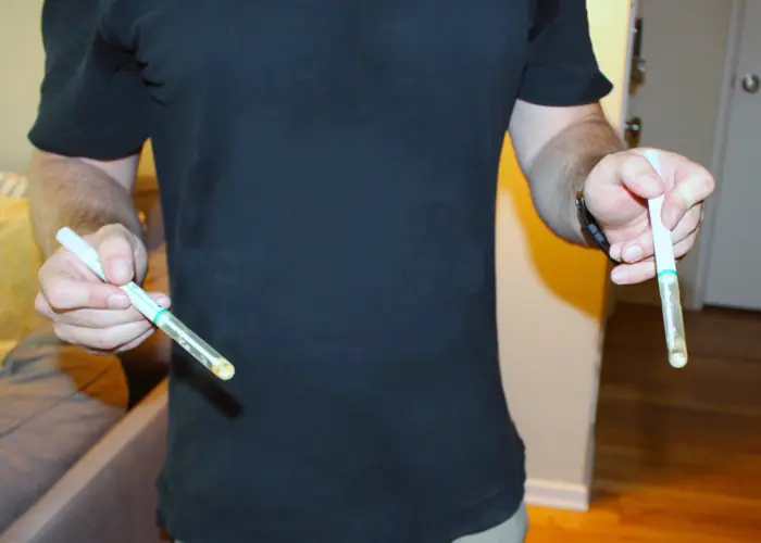 A person in a black shirt holds two test tubes, each containing a small, cloudy liquid, as they conduct mold testing indoors near a couch.