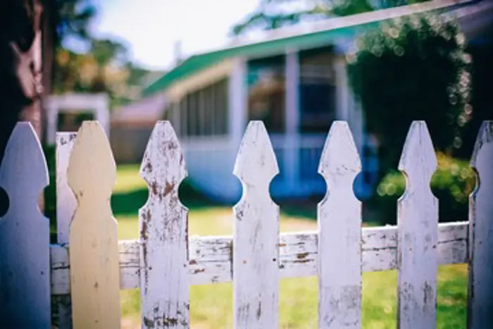 A close-up of a white picket fence with peeling paint suggests the need for an inspection. In the background, a bungalow with a screened porch and surrounding greenery is blurred. The scene conveys a sunny, rustic, suburban atmosphere.
