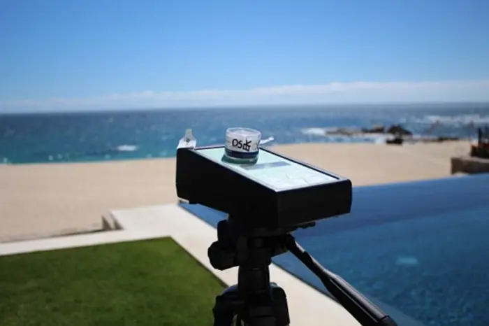 A device used for inspection is mounted on a tripod overlooking a beach with an infinity pool in the foreground. The sky is clear, and the ocean stretches in the background. A small container labeled Ds♂ rests atop the device, ready for sampling adventures ahead.