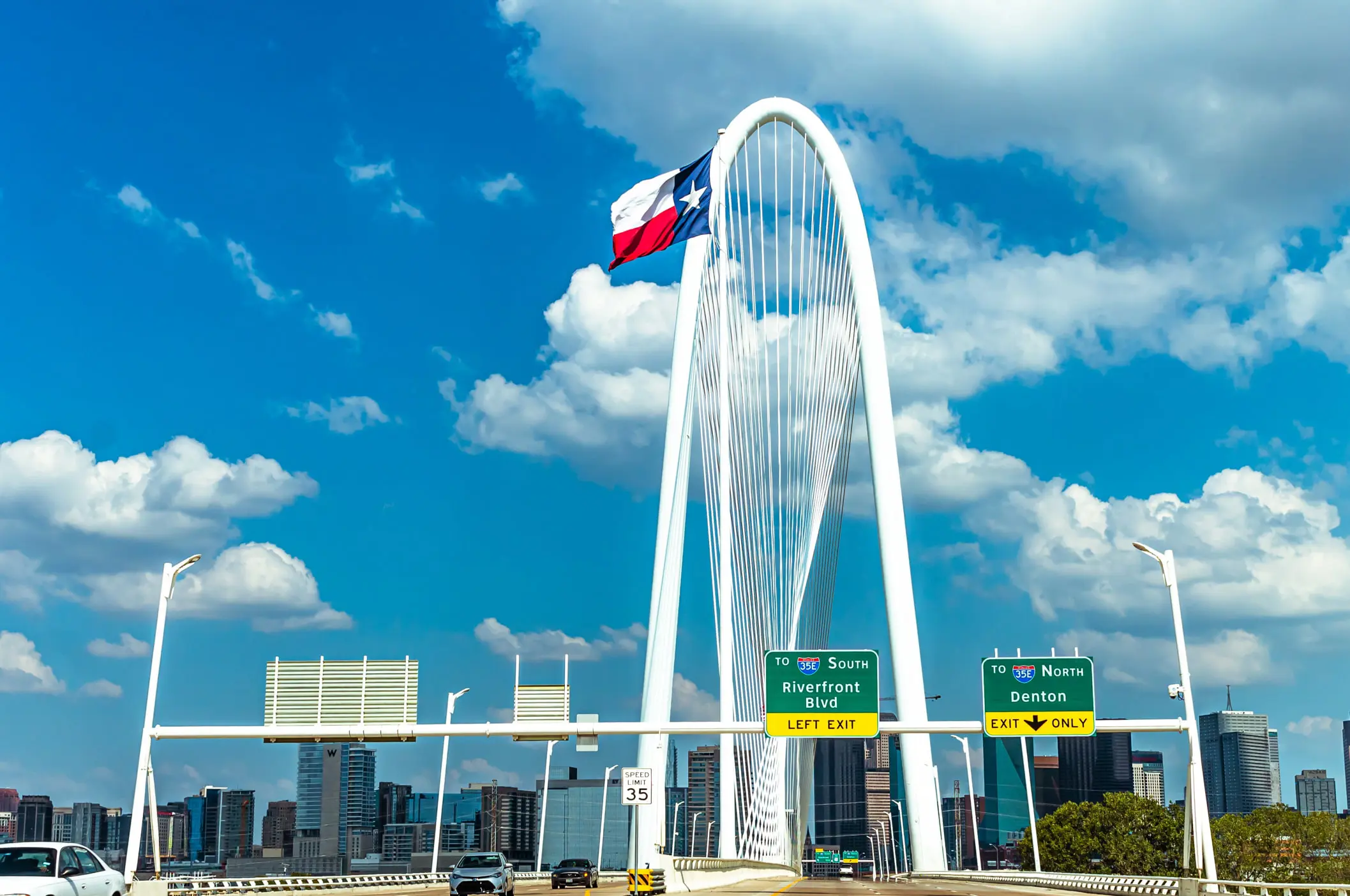 The image showcases the Margaret Hunt Hill Bridge in Dallas, Texas, where the Texas flag waves proudly against a blue sky. Road signs guide travelers to Riverfront Blvd and Denton, with the city's skyline providing a picturesque backdrop—an ideal scene for sampling urban landscapes.