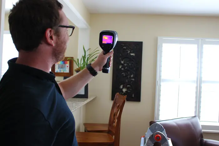 A man is conducting an inspection by holding and pointing a thermal camera towards a window in a room with a wooden chair and decorative wall art. The display on the camera reveals a detailed thermal image.