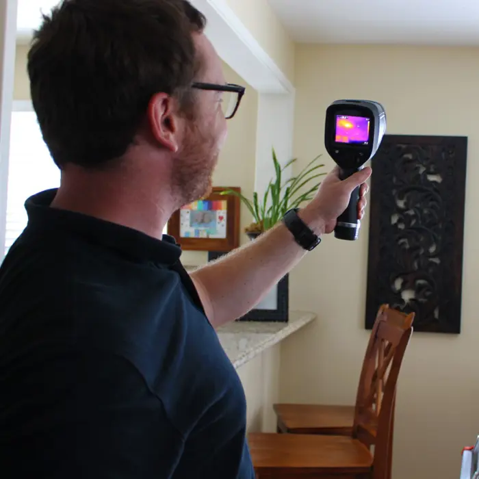 A person with short hair and glasses holds a thermal imaging camera indoors, conducting an inspection. The screen reveals a thermal color pattern. In the background are a wooden chair, decorative wall art, and a plant.