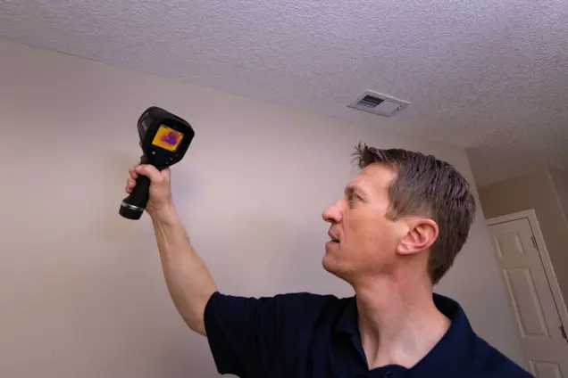 A man holding an infrared thermometer tests for temperature variations on a wall inside a house. The device displays a colored heat map, possibly indicating mold presence, and a ceiling vent is visible in the background.