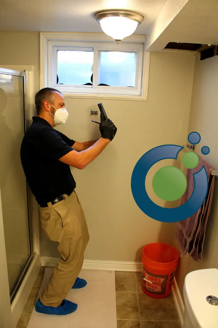 A person wearing a mask, gloves, and shoe covers is conducting a mold inspection on a bathroom wall with a toolbox on the floor. There's a circular logo with green and blue spheres on the wall as they meticulously photograph the area.