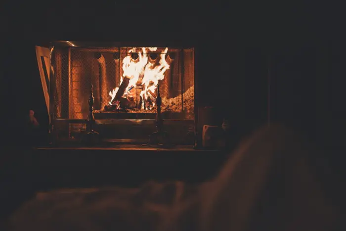 A warmly lit room features a glowing fireplace with burning logs. The flames cast a cozy ambiance as a blurred foreground suggests someone sampling the comfort nearby, wrapped in a blanket.