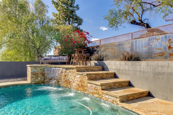 A tranquil backyard scene features a swimming pool with a small waterfall cascading over stone steps. Nearby, a wooden bar table with stools provides the perfect spot for assessment under a canopy of flowering bushes, surrounded by trees and a sunny, blue sky.