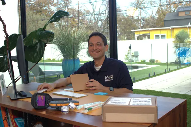 A person is sitting at a desk in a bright room with large windows, holding a small package, seemingly part of an ongoing investigation. The desk has various items, and outside is a yard with a pool and a yellow house. The person is smiling at the camera.