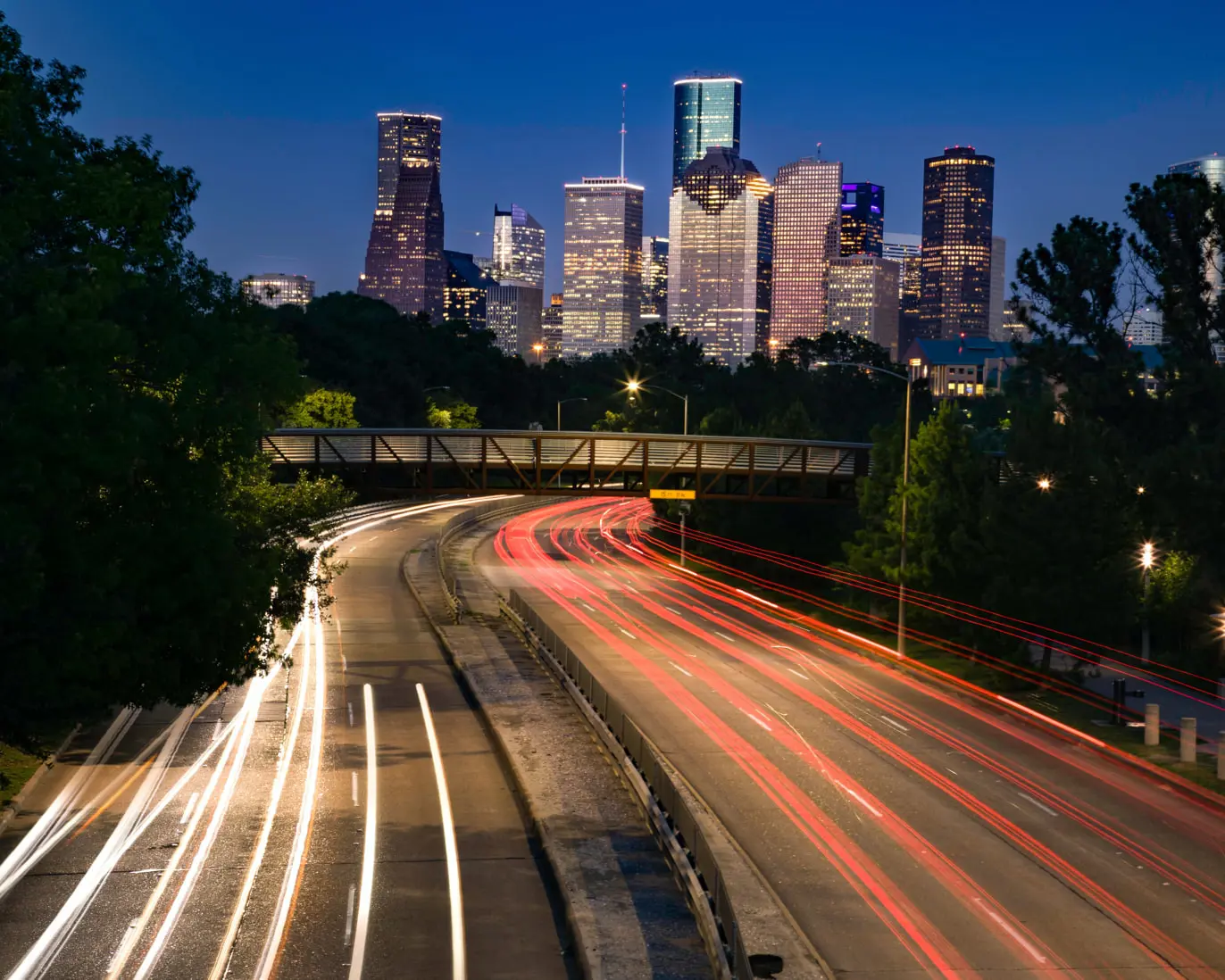 A long-exposure shot of a highway at night captures light trails from cars, resembling streaks of data sampling in motion. The city skyline, brightly illuminated, rises in the background under a clear blue sky. A pedestrian bridge spans the highway, while trees line the scene.