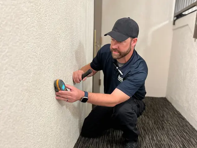 A person wearing a black cap and navy shirt kneels on a carpeted floor, using a handheld tool on a white wall. They hold a flashlight in the other hand, appearing to conduct an inspection or repair with meticulous precision.