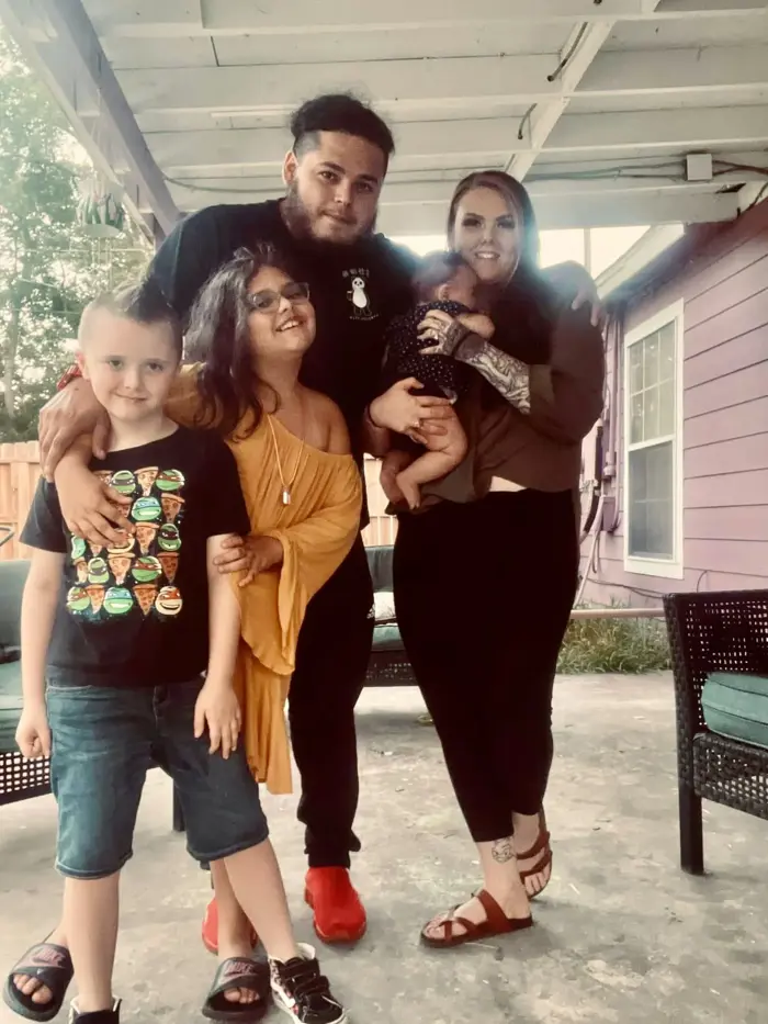 A family of five poses and smiles outside under a covered patio following a recent mold inspection. An adult man and woman stand in the back, with the woman holding a baby, while two children stand in front, dressed in yellow and black shirts.