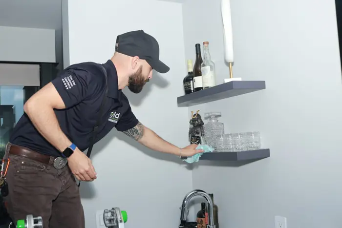 A man in a black cap and polo shirt is meticulously cleaning a shelf with a cloth, almost as if conducting an assessment. The shelf showcases several glass bottles and a stack of clear glasses, while a sculpture or white decorative piece sits elegantly on the top shelf.