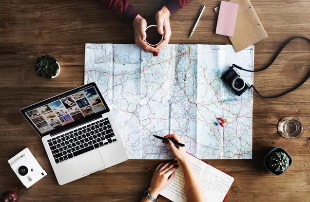 Two people are immersed in planning a trip at a wooden table. One conducts an assessment, marking their map with intent. Nearby, a laptop displays photos beside a camera, potted plant, notebooks, pen poised for notes, a cup in hand, and the square photo hinting at past adventures.