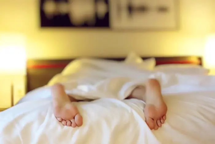 Two bare feet poke out from under a white blanket on a bed, with soft lighting from lamps on either side. The room has a warm, cozy ambiance, as if inviting an unhurried assessment of comfort, with a blurred wall picture in the background.
