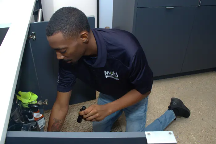 A man in a dark blue polo shirt and jeans is kneeling on the textured floor, conducting an inspection with a flashlight. He's checking for mold inside the cabinet, where cleaning supplies are visible. The room features dark cabinets, adding to the focused atmosphere of the task.