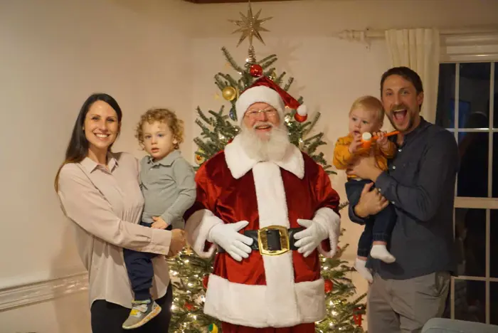 A happy family with two children poses with Santa Claus indoors, capturing a sampling of holiday joy. They stand in front of a decorated Christmas tree, with lights and ornaments. The parents hold the kids, who seem excited and cheerful. Santa is in the center, smiling warmly.