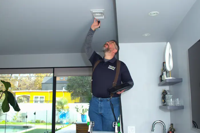 A maintenance worker, standing on a ladder, performs a mold assessment by adjusting a ceiling vent in a modern, bright room with large windows and a view of the backyard. He is holding a tool and appears focused on the task.