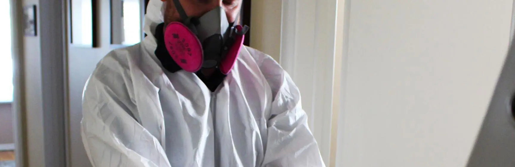 A person wearing a white protective suit, respirator mask with pink filters, and black gloves stands near an aluminum ladder in a hallway. The scene suggests preparation for inspection tasks requiring safety measures, such as cleaning or maintenance.