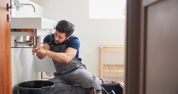 A plumber kneels on the floor, focused on fixing a pipe under a sink, as he conducts a routine inspection. Wearing gray overalls and a navy shirt, he is surrounded by his toolbox and a large black bucket. The room, with its beige walls and wooden bench, awaits his expert assessment.