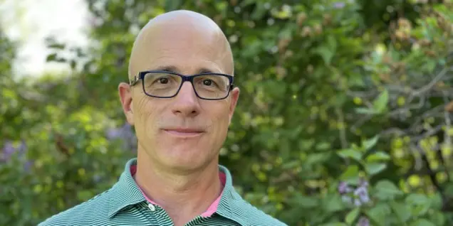 A person, looking poised for an investigation, wears glasses and a green and pink striped polo shirt as they stand in front of lush, leafy foliage.