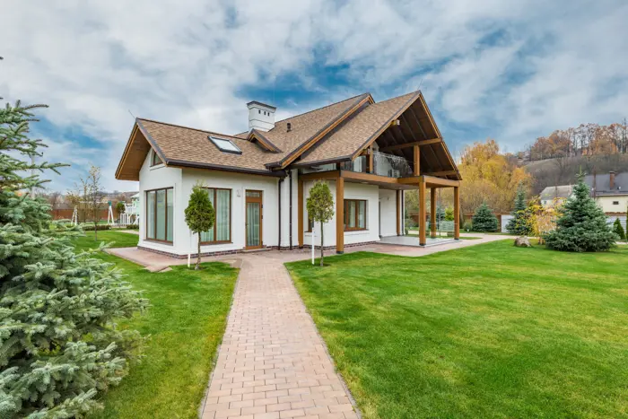 A modern, white house with a wooden roof is surrounded by a well-maintained lawn and paved pathways. The background features trees with autumn foliage under a partly cloudy sky, creating an ideal setting for an architectural assessment.
