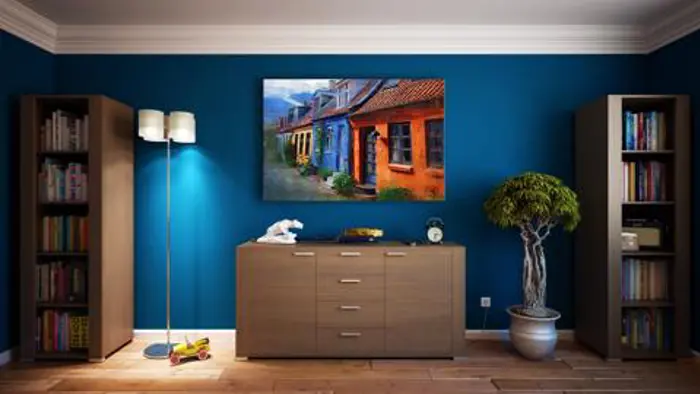 A modern living room with blue walls featuring a colorful street painting evokes an eye-catching assessment of style. There is a brown cabinet with decor items, flanked by two tall bookshelves. A potted plant stands to the right, and a floor lamp completes the balanced sampling on the left.