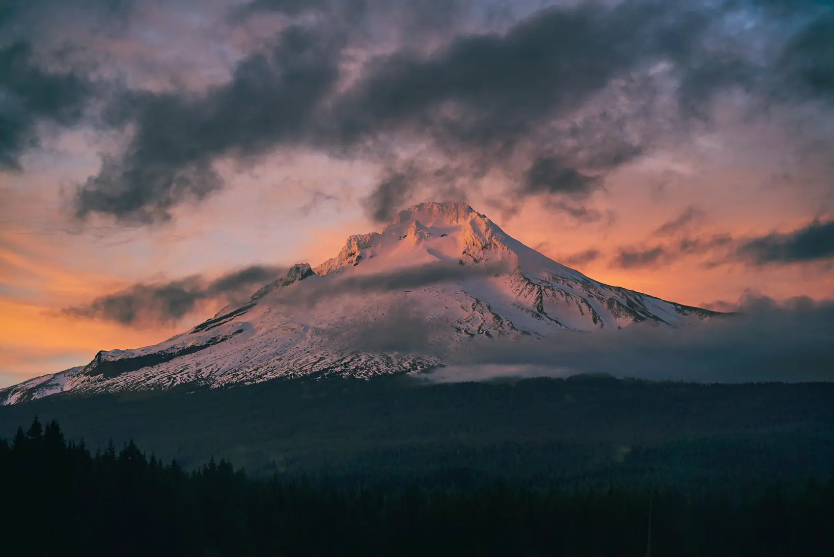 Snow-covered mountain peak under a dramatic, colorful sunset sky with clouds, as if nature is testing its own artistic limits. Forest silhouette in the foreground adds to the breathtaking scene.