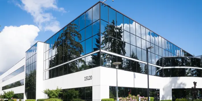 The modern glass office building at 15120 features reflective windows showcasing a clear blue sky and surrounding trees. With its sleek, minimalist design of white and glass, it invites a mold assessment to ensure the timeless beauty lasts on this sunny day.