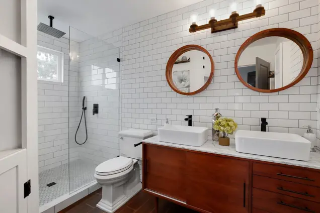 A modern bathroom features a glass-enclosed shower, white subway tile walls, and a wooden vanity with two round mirrors. Vessel sinks and black fixtures complete the look. A vase with flowers adds charm to the space, while recessed lighting illuminates it as if for an inspection.